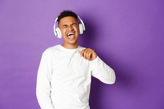 Image of carefree african-american guy dancing, listening music in wireless headphones and singing along, standing over purple background.