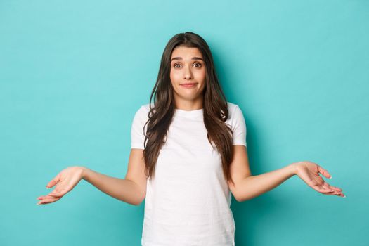 Image of pretty brunette woman in white t-shirt, dont know anything, shrugging and smiling clueless, standing over blue background.