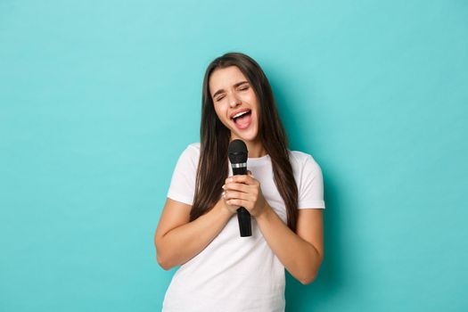Portrait of carefree brunette woman, having fun at karaoke bar, singing in microphone while standing over blue background.