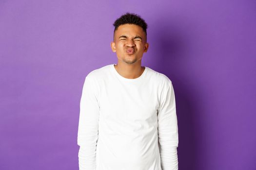 Image of silly and awkward african-american guy waiting for kiss, standing nervous with closed eyes over purple background.