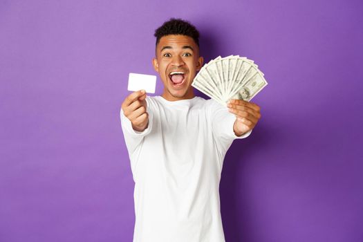 Image of handsome african american guy, looking pleased and smiling, showing credit card and money, standing over purple background.