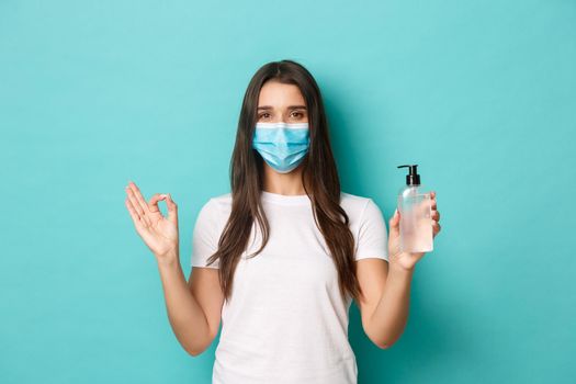 Concept of coronavirus, health and social distancing. Image of young brunette woman in medical mask, recommending to use hand sanitizer or antiseptic, showing okay sign.