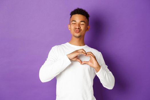 Image of lovely african-american boyfriend, waiting for kiss and showing heart sign, being in love, standing over purple background.