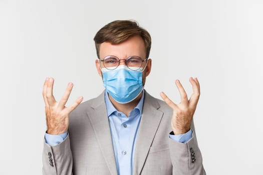 Concept of work, covid-19 and social distancing. Close-up of angry entrepreneur in gray suit and medical mask, shaking hands and grimacing mad or annoyed, standing over white background.