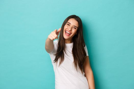Cheerful attractive woman in white t-shirt need you, pointing at camera and smiling, congratulating or picking someone, standing over blue background.