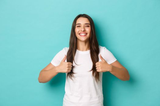 Image of satisfied smiling brunette girl, wearing white t-shirt, showing thumbs-up in approval, like and recommend product, standing over blue background.