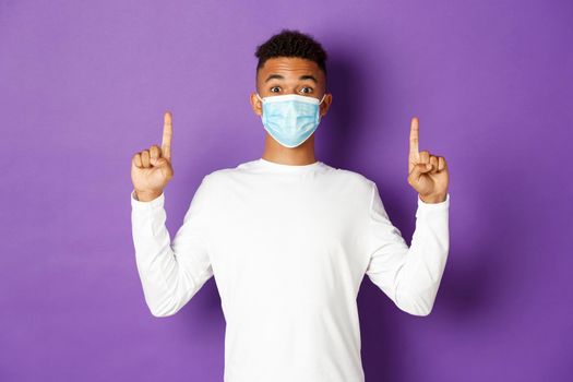 Concept of coronavirus, quarantine and lifestyle. Excited african-american guy in medical mask showing banner, pointing fingers up at copy space, standing over purple background.