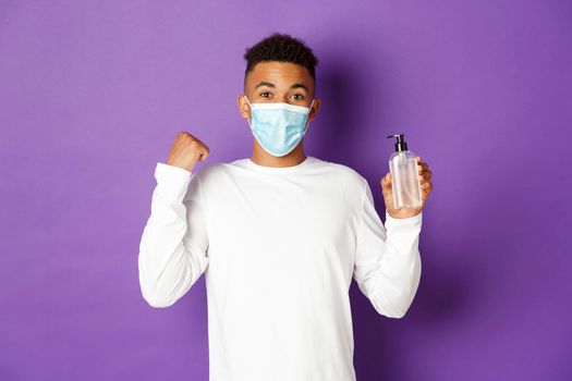 Concept of covid-19, pandemic and social distancing. Image of happy african-american man in medical mask, showing hand sanitizer and fist pump, rejoicing and standing over purple background.