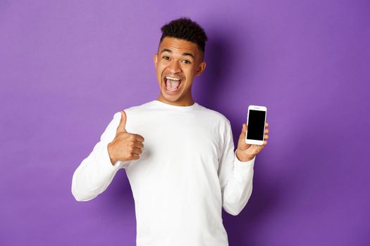 Image of african-american guy in white sweatshirt, smiling and showing thumbs-up in approval, showing mobile phone screen, standing over purple background.