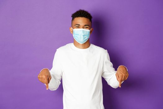 Concept of coronavirus, quarantine and lifestyle. Handsome african-american guy in medical mask showing promo, pointing fingers down at logo and smiling, standing over purple background.