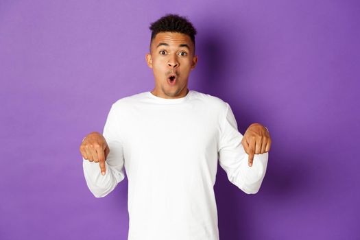 Image of handsome african-american man looking surprised, saying wow and pointing fingers down at something amazing, showing promo, standing over purple background.