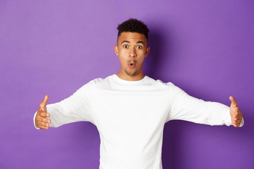 Amazed young african-american man showing something big, spread hands sideways to shape large object, standing over purple background.