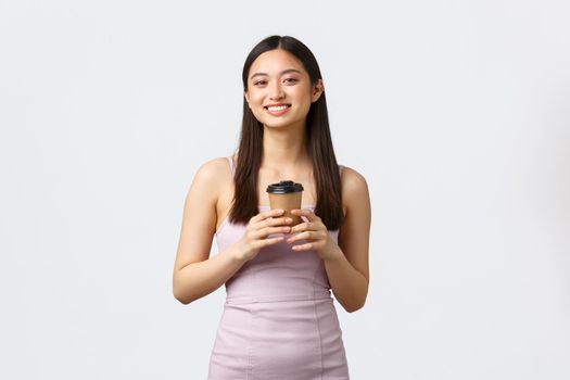 Lifestyle, people emotions and glamour concept. Beautiful young happy asian woman in dress, enjoying nice cup of coffee and smiling camera, order takeaway from cafe, white background.