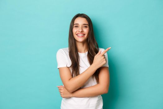 Cheerful brunette girl in white t-shirt, smiling pleased and pointing fingers at upper right corner, showing logo, standing over blue background.