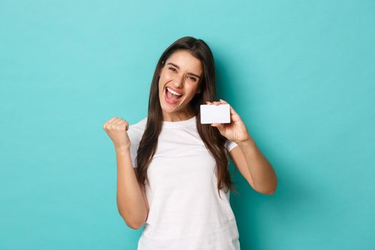 Image of satisfied ecstatic woman, wearing white t-shirt, rejoicing and showing credit card, standing pleased over blue background.