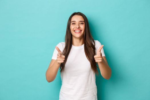 Cheerful brunette woman in white t-shirt, congratulating you, pointing fingers at camera, smiling pleased, praising good job, standing over blue background.