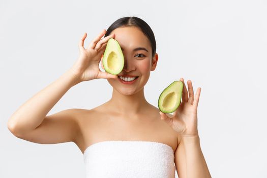 Beauty, personal care, spa and skincare concept. Close-up of tender feminine asian woman in bath towel, smiling and showing avocado near face, advertisement of face mask, cleanser or cream.