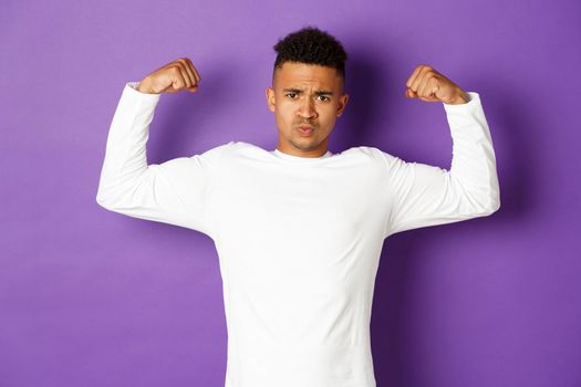 Image of confident african-american young guy, flexing biceps and trying to show-off, showing muscles after workout, standing over purple background, feeling strong.