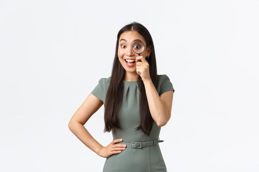 Small business owners, women entrepreneurs concept. Excited happy asian woman found something good, looking through magnifying glass with pleased smile, searching for product, white background.