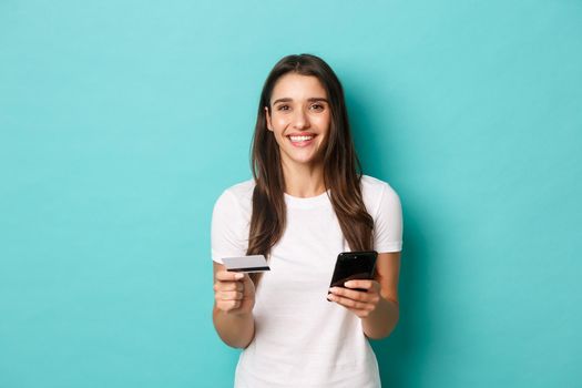 Portrait of young brunette woman in casual white t-shirt, shopping online, holding credit card and mobile phone, standing over blue background.