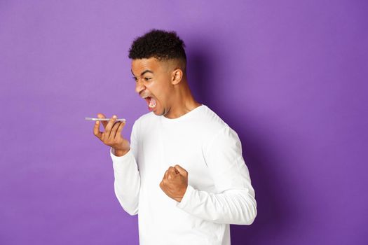 Image of angry african-american man shouting at speakerphone, looking angry and pissed-off, standing with mobile phone over purple background.