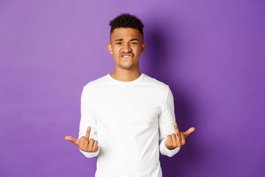 Image of arrogant and rude african-american guy in white sweatshirt, showing middle-finger, fuck you signs and grimacing, standing over purple background.