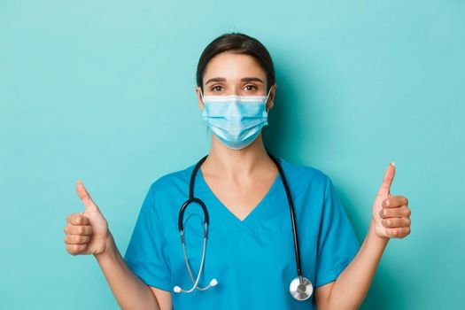 Concept of covid-19 and quarantine concept. Close-up of confident beautiful female doctor in scrubs and medical mask, guarantee safety, showing thumbs-up, standing over blue background.