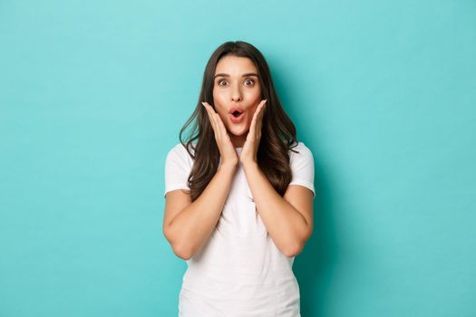 Image of surprised brunette girl reacting to announcement, gasping and looking at camera fascinated, gossiping over blue background.