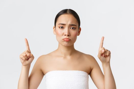 Beauty, cosmetology and spa salon concept. Disappointed pouting asian girl in white towel complaining or being jealous, pointing fingers up and whining about promo, white background.