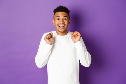 Image of hopeful african-american man, looking with anticipation at camera, smiling and jumping from excitement, standing over purple background.