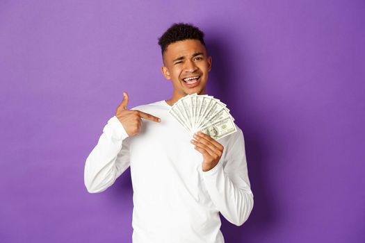 Image of sassy african-american guy smiling, bragging with big sum of money, pointing at cash, standing over purple background.