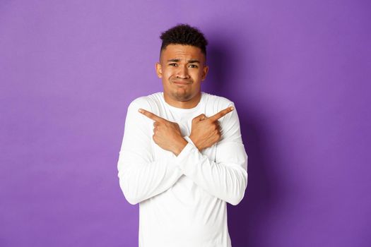 Image of indecisive african-american guy, uncomfortable making decision, asking for help with choice, pointing fingers sideways at left and right copy space, standing over purple background.