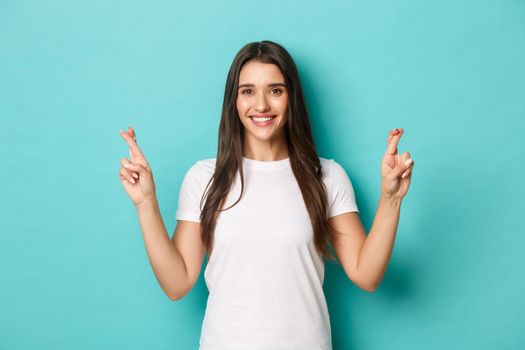 Confident attractive woman, looking hopeful and smiling, crossing fingers for good luck, making wish, standing over blue background.