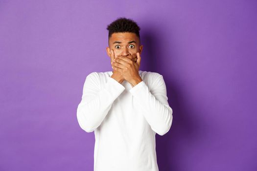 Image of scared and shocked african-american man, reacting to something frightening, gasping and cover mouth with hands, looking at camera, standing over purple background.