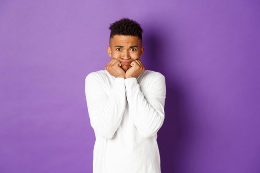 Image of insecure and scared african-american man, trembling from fear and looking at something scary, standing over purple background in white sweatshirt.