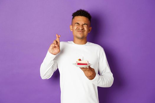 Image of excited african-american man celebrating birthday, making wish, holding b-day cake and crossing fingers, standing over purple background.