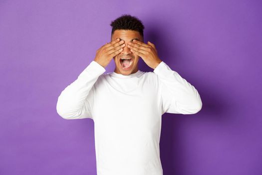 Image of handsome african-american 20s guy, cover eyes with hands and smiling, waiting for surprise with excited face, standing over purple background.