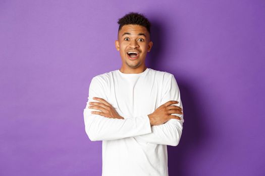 Image of handsome young african-american guy in white sweatshirt, open mouth and looking surprised, standing near purple copy space for your logo.