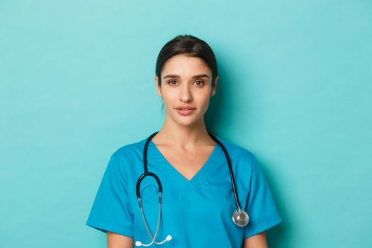 Concept of covid-19 and quarantine concept. Close-up of young confident female doctor with stethoscope, wearing scrubs, standing over blue background.