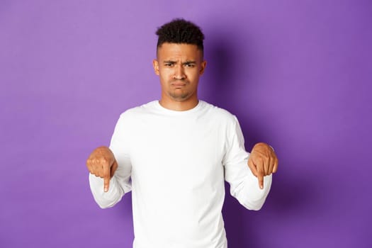 Portrait of silly upset african-american guy, frowning and sulking, complaining, pointing fingers down at something disappointed, standing over purple background.