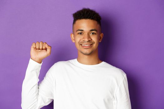 Close-up of handsome smiling african-american man, looking hopeful and raising fist, showing black lives matter sign, fighting for rights, standing over purple background.