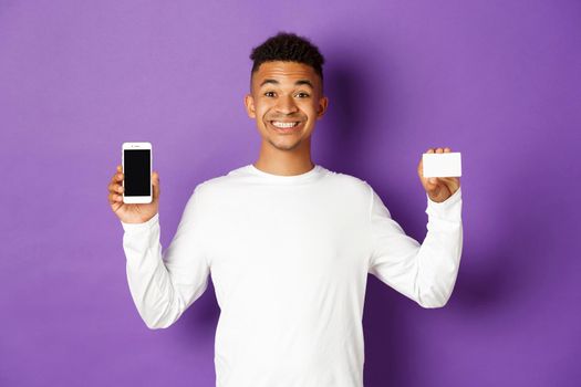 Portrait of handsome african-american man, smiling pleased, showing smartphone screen and credit card, standing over purple background.