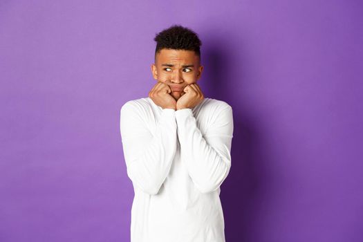 Image of insecure and scared african-american man, trembling from fear and looking left at something scary, standing over purple background.
