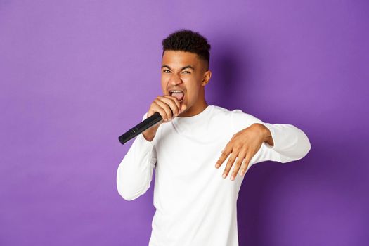 Image of sassy african-american guy singing in microphone, rapping and performing on stage, standing over purple background.