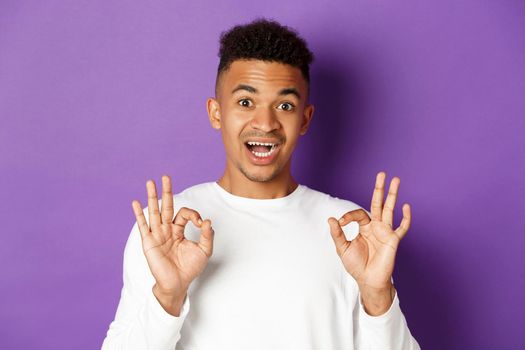 Close-up of handsome young african-american man, looking amazed and showing okay signs, recommend something good, standing over purple background.