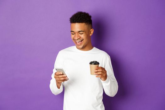 Happy african-american man drinking coffee, laughing and looking at smartphone, standing over purple background.