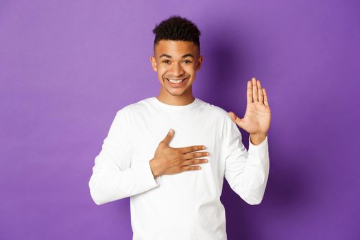 Image of handsome young african-american guy, being honest, holding arm raised and hand on heart, promise something or swear to tell truth, standing over purple background.
