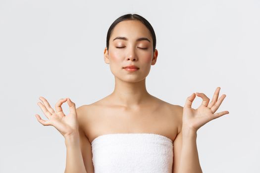 Beauty, cosmetology and spa salon concept. Close-up of meditating relaxed beautiful asian female in white towel make zen gesture with closed eyes, feeling pleased after massage therapy.