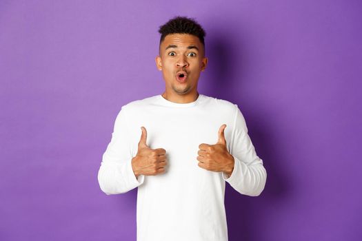 Impressed handsome african-american guy, showing thumbs-up in approval, praise great promo, like and recommend, looking amazed at camera, standing over purple background.