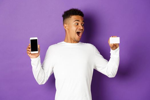 Portrait of handsome african-american man, looking amazed at credit card and showing app on mobile phone screen, standing over purple background.